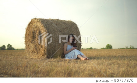 A young woman sits near a haystack in a field with a phone in her hands. A girl with glasses and a blue dress is texting on the phone. 90757870