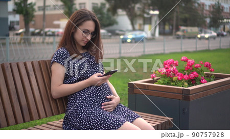Young pregnant girl with a phone in her hands. A girl with glasses and a dress is texting on the phone. 90757928