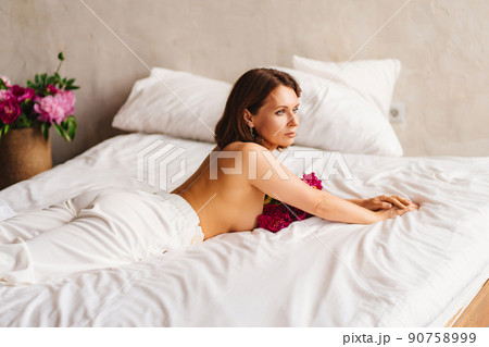 attractive brunette topless woman with a bouquet of peonies on the bed. 90758999