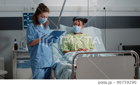 Medical assistant doing consultation with ill patient, wearing face masks in hospital ward. Nurse discussing with young woman about healthcare treatment and taking notes during pandemic 90759290