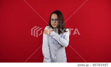 A young woman in glasses has a sore shoulder and she touches it with her hand. Shooting on a red background. A young woman in glasses has a sore shoulder and she touches it with her hand. Shooting on a red background. 90765497