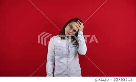 A young woman with glasses has a headache and holds it in her hands. Shooting on a red background. 90765515