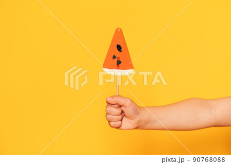 Child's hand holding lollipop in the shape of piece of watermelon against yellow background 90768088