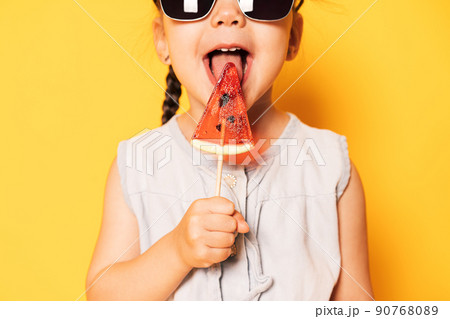 Girl in sunglasses licks lollipop in the form of piece of watermelon against yellow background 90768089