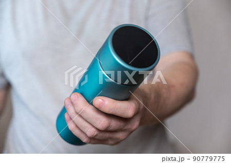 Middle section of man holding vacuum flask in his hand. Adult man holding cylindrical turquoise thermos with lid. Container for hot and cold drinks. Equipment for sports, travel, hiking and tourism. 90779775