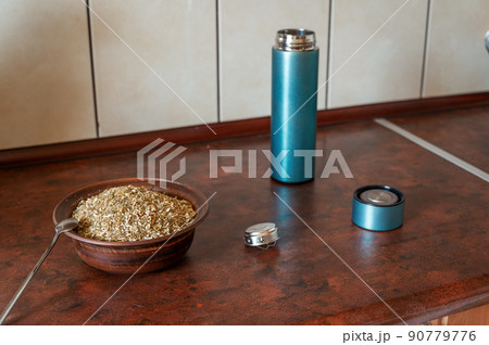 Dried Matricaria chamomilla flowers in a bowl on the kitchen table. An open empty cylindrical thermos with a lid and a teapot next to it. The process of making an herbal tea or infusion of chamomile. 90779776