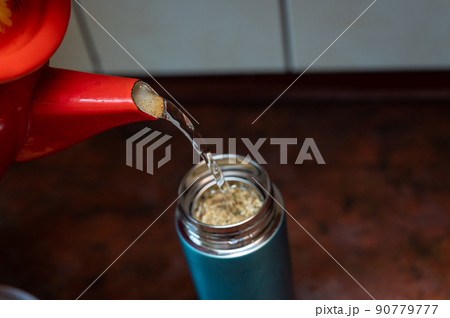 Boiling water from the red chanter is poured into a thermos. The teapot is filled with medicinal herbs. Dried flowers Matricaria chamomilla. Selective focus. Series part. Boiling water from the red chanter is poured into a thermos. The teapot is filled with medicinal herbs. Dried flowers Matricaria chamomilla. Selective focus. Series part. 90779777