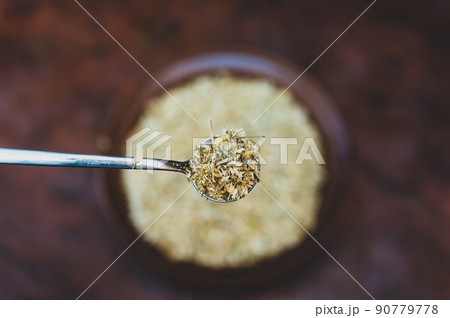 Full spoonful of dried Matricaria chamomilla flowers above the kitchen table. Brown bowl of medicinal herbs blurred in the background. Process of making an herbal tea or infusion of chamomile. 90779778