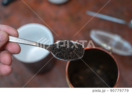 Middle-aged man holding spoonful of dry tea. Crushed black tea leaves in spoon over red table. Cup and container are blurred on background. Healthy Lifestyle. First-person view. Front focus. Indoor. Middle-aged man holding spoonful of dry tea. Crushed black tea leaves in spoon over red table. Cup and container are blurred on background. Healthy Lifestyle. First-person view. Front focus. Indoor. 90779779