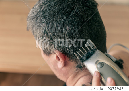 Wife cuts her husband's hair at home. Woman uses an electric hair clipper to cut man's hair at back of his head. Middle-aged person with naked torso sits inside living room. Selective Focus. Wife cuts her husband's hair at home. Woman uses an electric hair clipper to cut man's hair at back of his head. Middle-aged person with naked torso sits inside living room. Selective Focus. 90779824