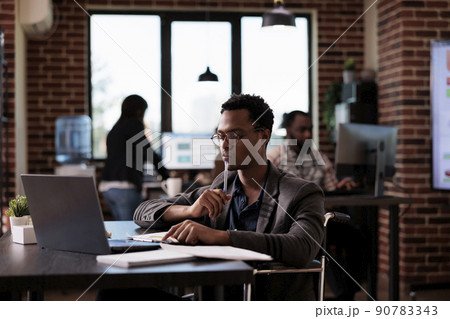 Male wheelchair user with chronic health condition working on laptop website, reading email at desk. Paralyzed company employee doing presentation in disability friendly office. 90783343