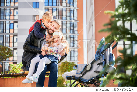 Cheerful family hugging together, spending time outdoors while baby sleeping in pram. Happy father sitting on bench with son and daughter at modern courtyard of city residential high-rise buildings. 90785017