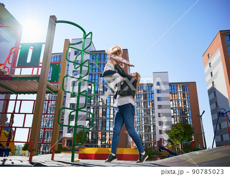 Happy blond mom catching cheerful daughter, jumping from ladder at playground. Beautiful woman having fun with female child at modern courtyard of city residential high-rise buildings. Happy blond mom catching cheerful daughter, jumping from ladder at playground. Beautiful woman having fun with female child at modern courtyard of city residential high-rise buildings. 90785023