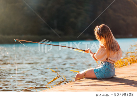 Summer hobby. A young Caucasian blonde woman is fishing on a wooden pier. Back view. The concept of summer vacation 90799308