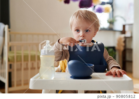 Cute blonde little boy in bib eating porridge with spoon sitting in his chair Cute blonde little boy in bib eating porridge with spoon sitting in his chair 90805812