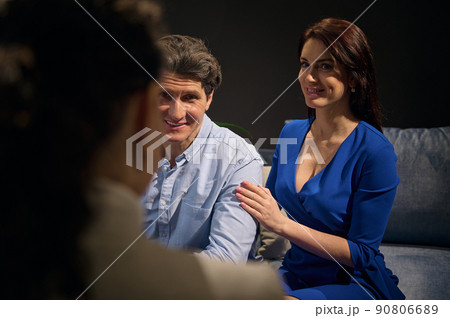 Beautiful Caucasian woman in casual blue dress sitting on a comfy sofa next to her husband and talking with a blurred woman on the foreground Beautiful Caucasian woman in casual blue dress sitting on a comfy sofa next to her husband and talking with a blurred woman on the foreground 90806689