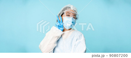 Happy, kawaii asian female doctor, nurse in personal protective equipment, showing v-sign peace gesture near face mask, blue background Happy, kawaii asian female doctor, nurse in personal protective equipment, showing v-sign peace gesture near face mask, blue background 90807200