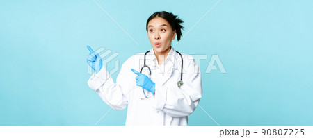 Surprised asian woman doctor or nurse, pointing fingers left and looking amazed at advertisement, copy space, standing in uniform over blue background Surprised asian woman doctor or nurse, pointing fingers left and looking amazed at advertisement, copy space, standing in uniform over blue background 90807225