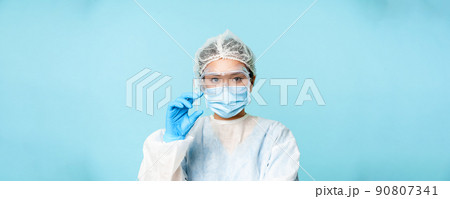 Close up portrait of asian female doctor, nurse in personal protective equipment showing syringe with vaccine, vaccinating from flu or covid, blue background 90807341