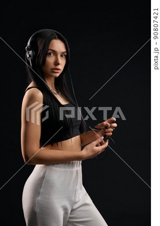 Gentle dark haired lady wearing top, listening music with headphones. Side view of attractive sporty girl in white sweatpants enjoying song with earphones, black background. Concept of lifestyle. Gentle dark haired lady wearing top, listening music with headphones. Side view of attractive sporty girl in white sweatpants enjoying song with earphones, black background. Concept of lifestyle. 90807421