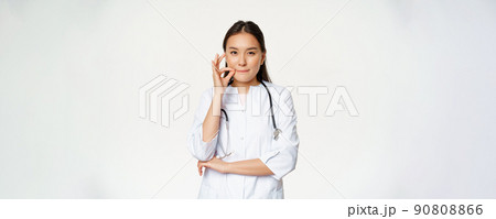 Physician-patient privilege, medical confidentiality. Asian woman doctor showing silence, mouth zip gesture, secret, standing over white background Physician-patient privilege, medical confidentiality. Asian woman doctor showing silence, mouth zip gesture, secret, standing over white background 90808866
