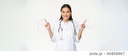 Smiling professional physician in white robe, pointing fingers sideways and showing two advertisements, both ways, standing against white background 90808867