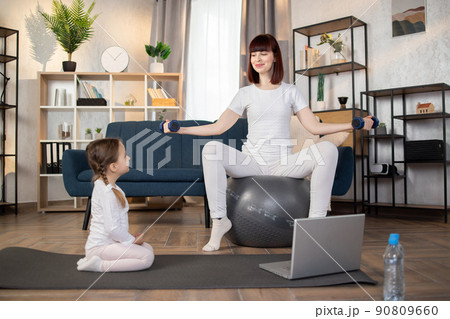 Woman doing sports sitting on fitball with dumbbells in hands while herdaughter sitting on mat 90809660