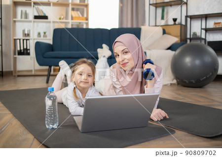 Smiling young muslim mother lying with her cute little daughter on mat on living room floor at home 90809702