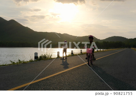 Cute little girl riding a bicycle and her sister riding a scooter on a lake road at sunset. Happy sisters doing outdoor activities together. Healthy Summer Activities for Kids. Cute little girl riding a bicycle and her sister riding a scooter on a lake road at sunset. Happy sisters doing outdoor activities together. Healthy Summer Activities for Kids. 90821755