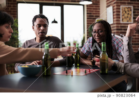 Multiethnic group of friends sitting at table while playing boardgames with figurines. Happy diverse people enjoying society games together while having snacks and beverages. 90823064