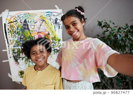 Two kids smiling at camera of smartphone while making selfie portrait against the artwork on wall Two kids smiling at camera of smartphone while making selfie portrait against the artwork on wall 90827477