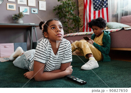 African girl lying on floor and watching movie on TV with her little brother playing smartphone in background African girl lying on floor and watching movie on TV with her little brother playing smartphone in background 90827650