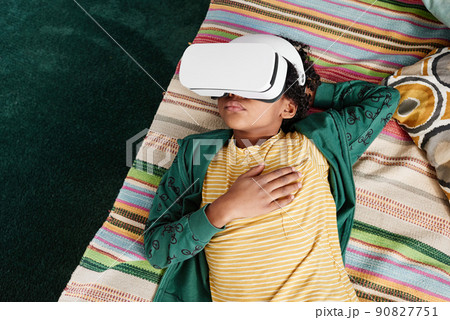 High angle view of African little boy relaxing on sofa and using VR goggles to watch virtual reality video during his leisure time at home 90827751