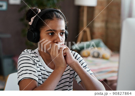 African serious girl in wireless headphones sitting in front of computer at home and looking at monitor 90827864