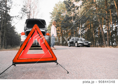 Red emergency triangle with blurred car and woman calling car mechanic in the background. Accident and broken car on the road Red emergency triangle with blurred car and woman calling car mechanic in the background. Accident and broken car on the road 90844348