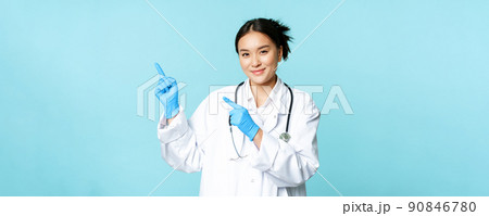 Smiling female doctor, physican in medical uniform and sterile gloves, pointing fingers left at promo, clinic logo, standing over blue background 90846780
