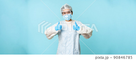 Optimistic asian nurse or doctor, wearing personal protective equipment, showing thumbs up, campaign from coronavirus prevention, blue background 90846785