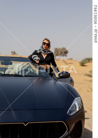 A stylish girl in a black suit and a silk scarf enjoys a ride in a convertible car in the desert, Dubai, UAE 90861962