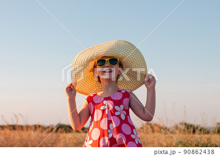 Child girl in straw hat dress in wheat field. Smiling kid in sunglasses sunset countryside. Cottagecore style aesthetic. 90862438