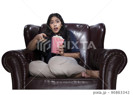 Asian woman holding a television remote and popcorn sitting on the couch with a scared expression Asian woman holding a television remote and popcorn sitting on the couch with a scared expression 90863342