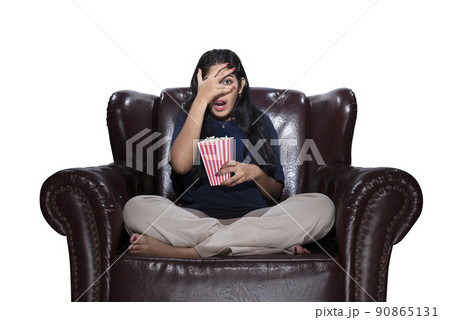 Asian woman with popcorn sitting on the couch with a scared expression Asian woman with popcorn sitting on the couch with a scared expression 90865131