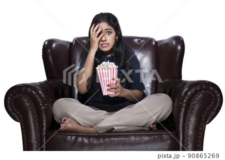 Asian woman with popcorn sitting on the couch with a scared expression 90865269