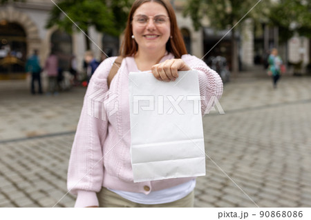Young happy redhead woman in glasses holding take away paper bag over street background. White Packaging template mock up. Delivery service concept. Blank snack white paper bag Young happy redhead woman in glasses holding take away paper bag over street background. White Packaging template mock up. Delivery service concept. Blank snack white paper bag 90868086