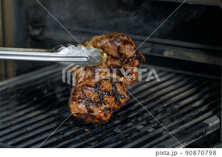 Silver serving kitchen tongs holding meat beef steak grilling on rack charcoal stove with smoke on black background. 90870798