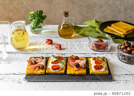 Grilled polenta with stracchino cheese, tomatoes, herbs, speck and olive. White wooden background, ingredients and table wine. Grilled polenta with stracchino cheese, tomatoes, herbs, speck and olive. White wooden background, ingredients and table wine. 90871202
