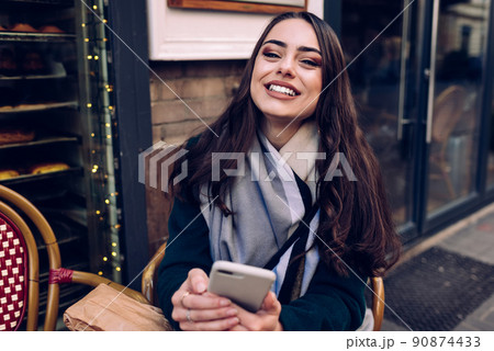 Cheerful young woman using smartphone near entrance of pastry shop 90874433