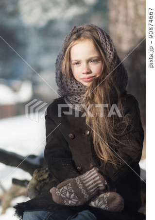 cute caucasian girl wearing warm coat, wool mittens and shawl sitting by the tree on cold winter day. cute caucasian girl wearing warm coat, wool mittens and shawl sitting by the tree on cold winter day. 90874667