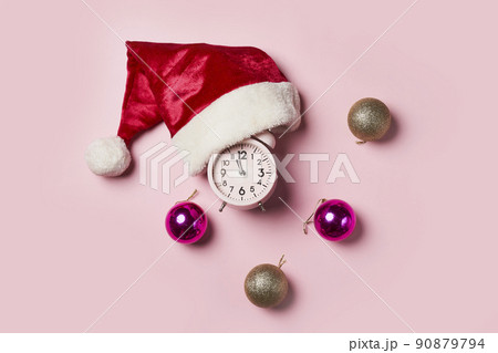 Christmas time consept.Christmas clock in a Santa Claus hat or cap on a pink background. The minute hand at twelve o'clock on New Year's Eve. Christmas time consept.Christmas clock in a Santa Claus hat or cap on a pink background. The minute hand at twelve o'clock on New Year's Eve. 90879794