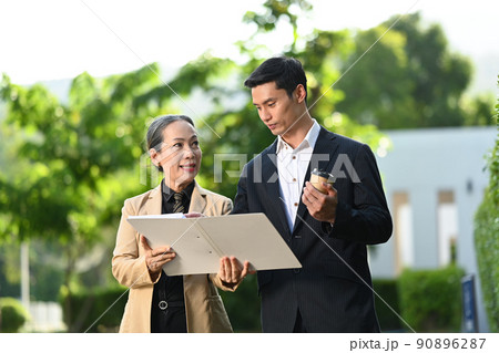 Attractive mature female manager and young businessman talking each other while walking outside modern office building Attractive mature female manager and young businessman talking each other while walking outside modern office building 90896287