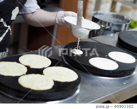The guy cook in gloves pours a portion of batter with a ladle on an electric stove for baking pancakes. Fresh pastries. 90901302